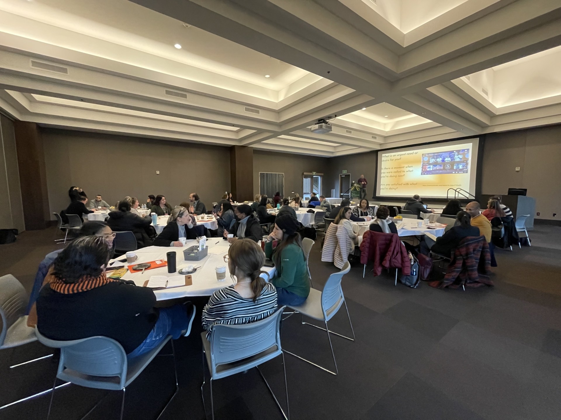 Conference room filled with people sitting at circle tables with a presentation on a screen.