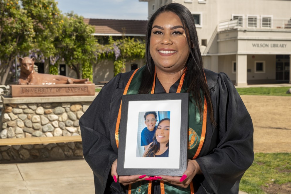 Janelle Bowens. graduating student of 2023, standing in front of the Leopard statue on campus in her regalia holding an image of her and her son.
