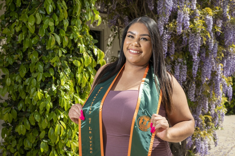 Janelle Bowens is a graduate, standing in front of a puple flower bush with sash