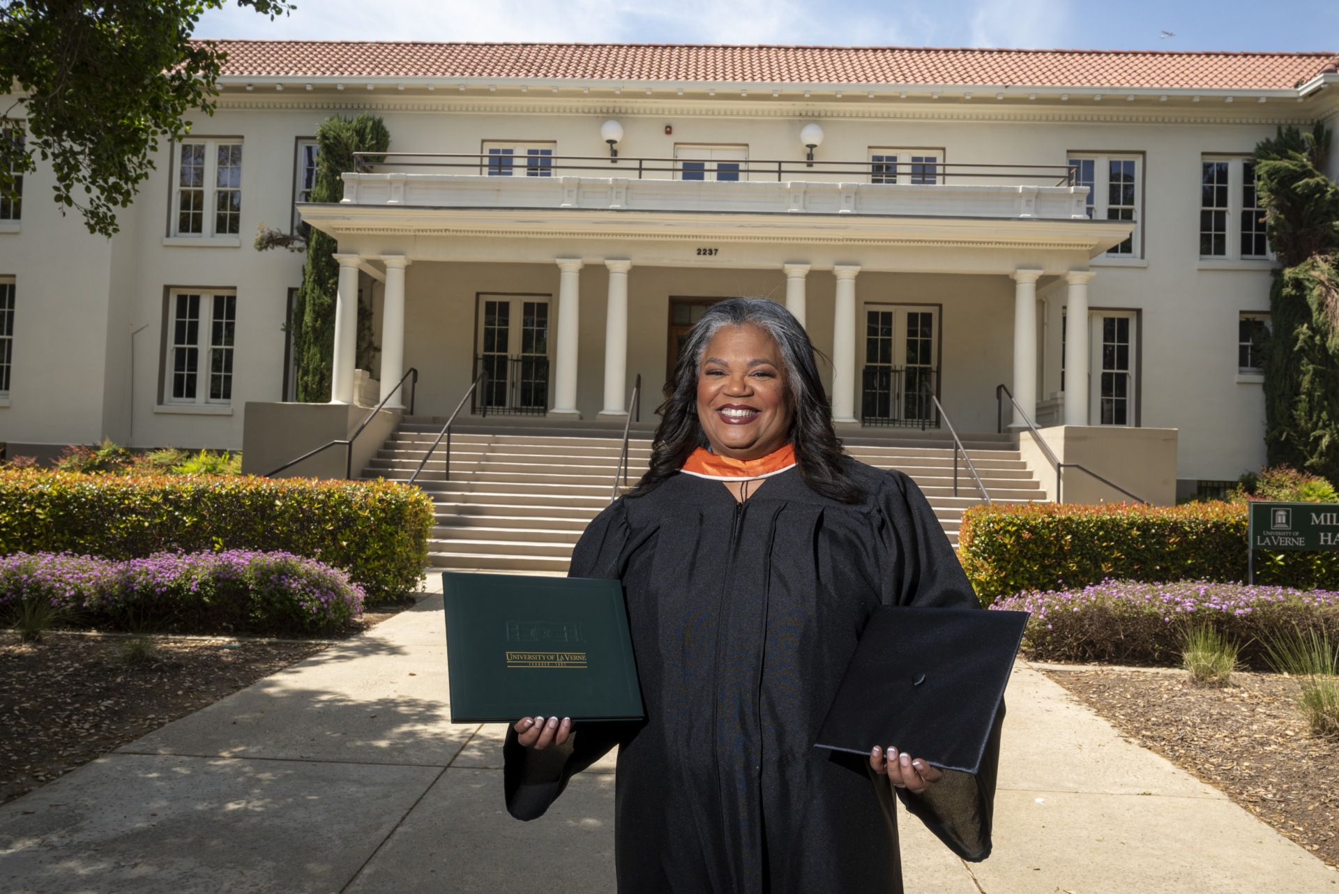 Darcelle Jones-Wesley, 2023 grad, stands in her regalia in front of Miller Hall with a diploma and her cap in her hands.