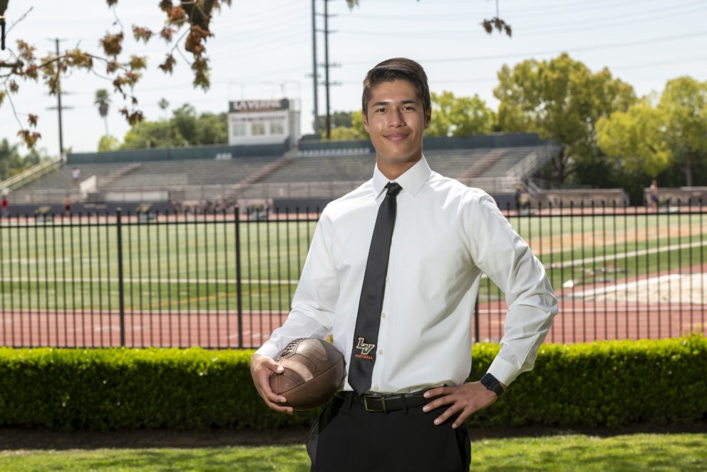 2023 graduate Kapena stands in front of Ortmayer Stadium holding football in suit