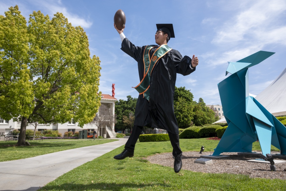 Student grad Kapena jumps for the football in his regalia