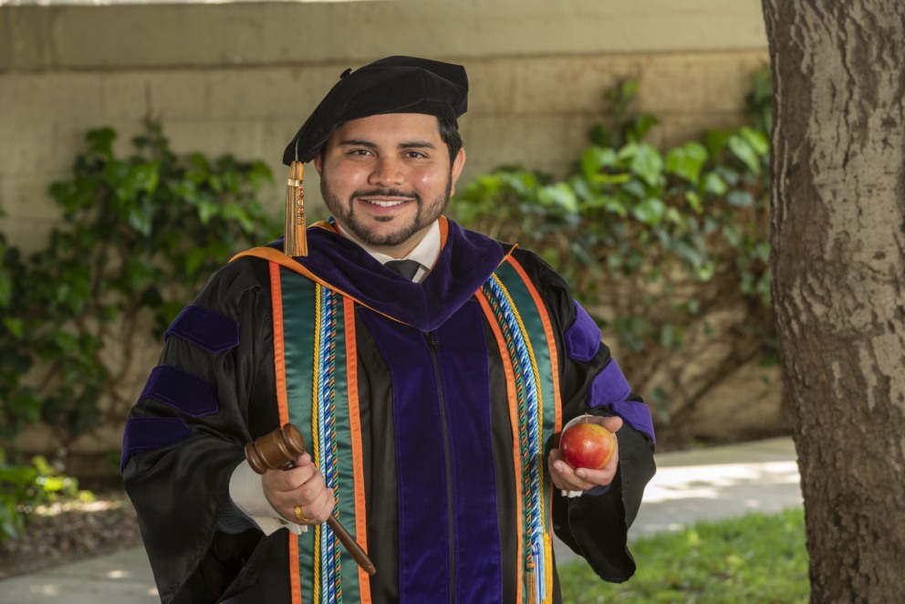 Jeffrey Torres, graduate of 2023, stands with a gavel and an apple outside in his regalia